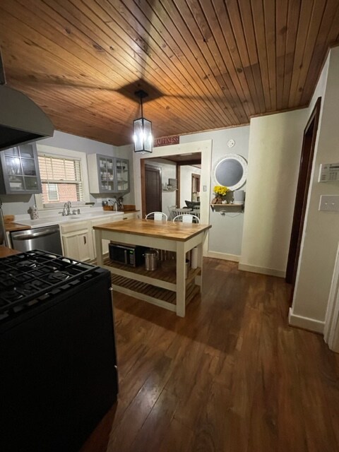 The kitchen features wood-paneled ceilings and a central island with shelves. Stainless steel appliances are present along with white cabinetry. A window allows natural light, while a small dining area is visible in the background. Decorative elements include flowers and a round mirror.