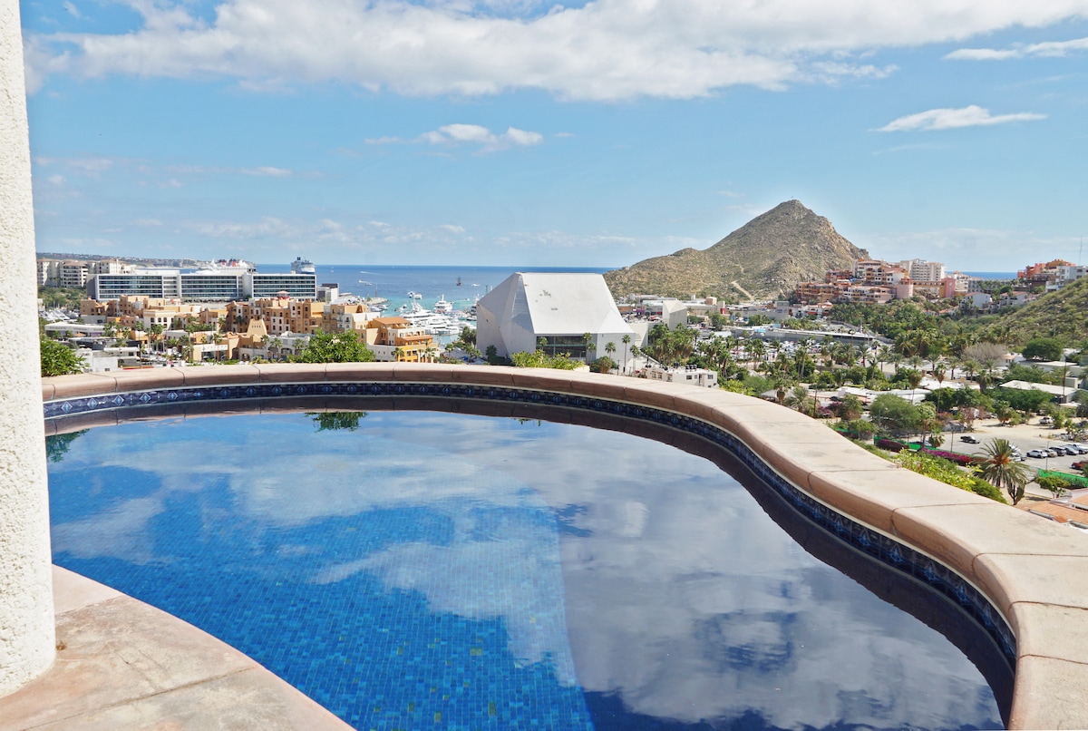 An infinity-edge pool reflects the sky, seamlessly blending with the horizon. The view captures the marina and coastline of Cabo San Lucas, framed by gentle hills. Lush greenery and scattered buildings are visible in the distance, enhancing the scenic backdrop.