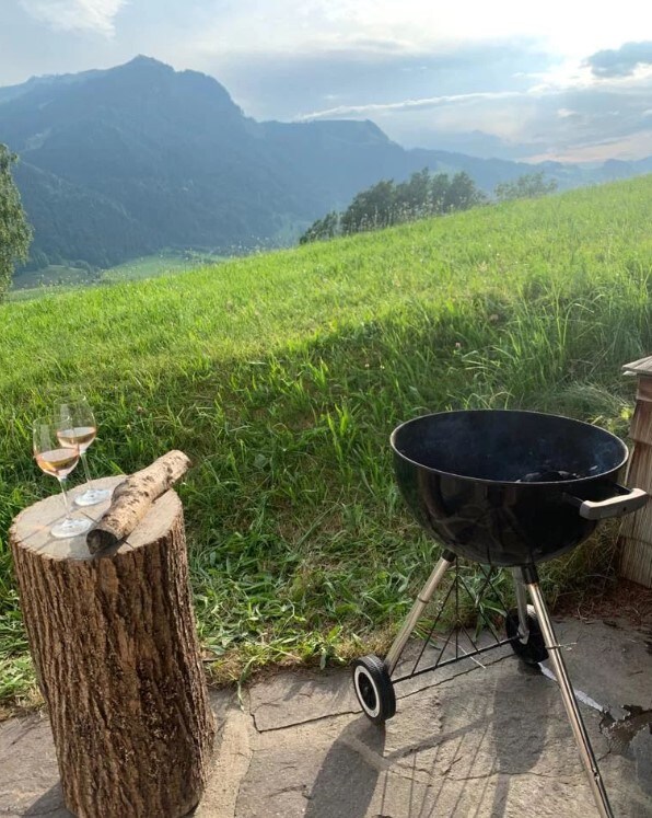 A charcoal grill stands on a stone patio, accompanied by two glasses of rosé wine on a wooden stump. Lush green grass stretches out toward the mountains, under a serene sky with soft clouds.