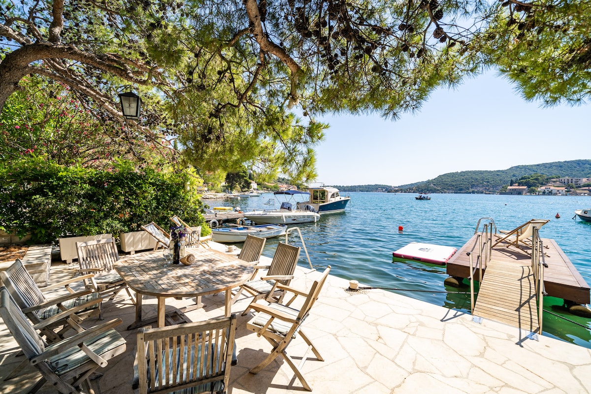 An outdoor patio area features a wooden table surrounded by several chairs, shaded by a large tree. A view of the water is visible, along with boats moored nearby. A private dock extends into the water, providing access for swimming or boating.
