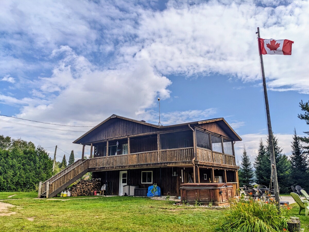 The chalet features a spacious, two-story wooden structure with a large outdoor deck and screened porch. A Canadian flag gently flutters in the breeze. Lush greenery and tall trees surround the property, offering a sense of seclusion.