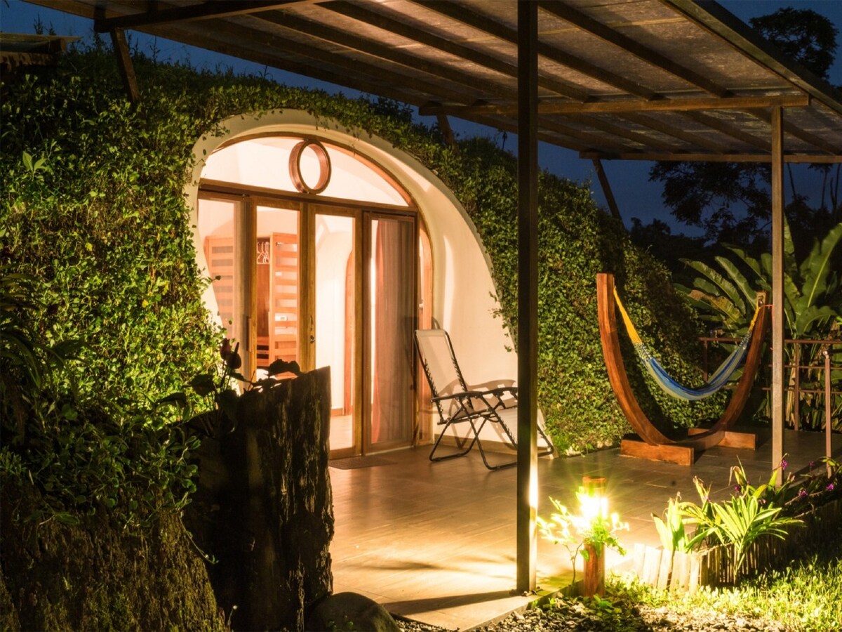 An inviting entrance is illuminated by soft lighting at night, showcasing an arched doorway framed by lush greenery. A comfortable hammock is hanging nearby, while a chair sits on the covered patio, inviting relaxation in a serene outdoor setting.