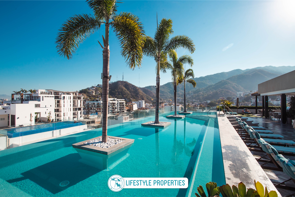 A serene rooftop infinity pool is framed by palm trees, offering panoramic views of the mountains and surrounding area. Comfortable lounge chairs line the deck, inviting relaxation under the clear blue sky.
