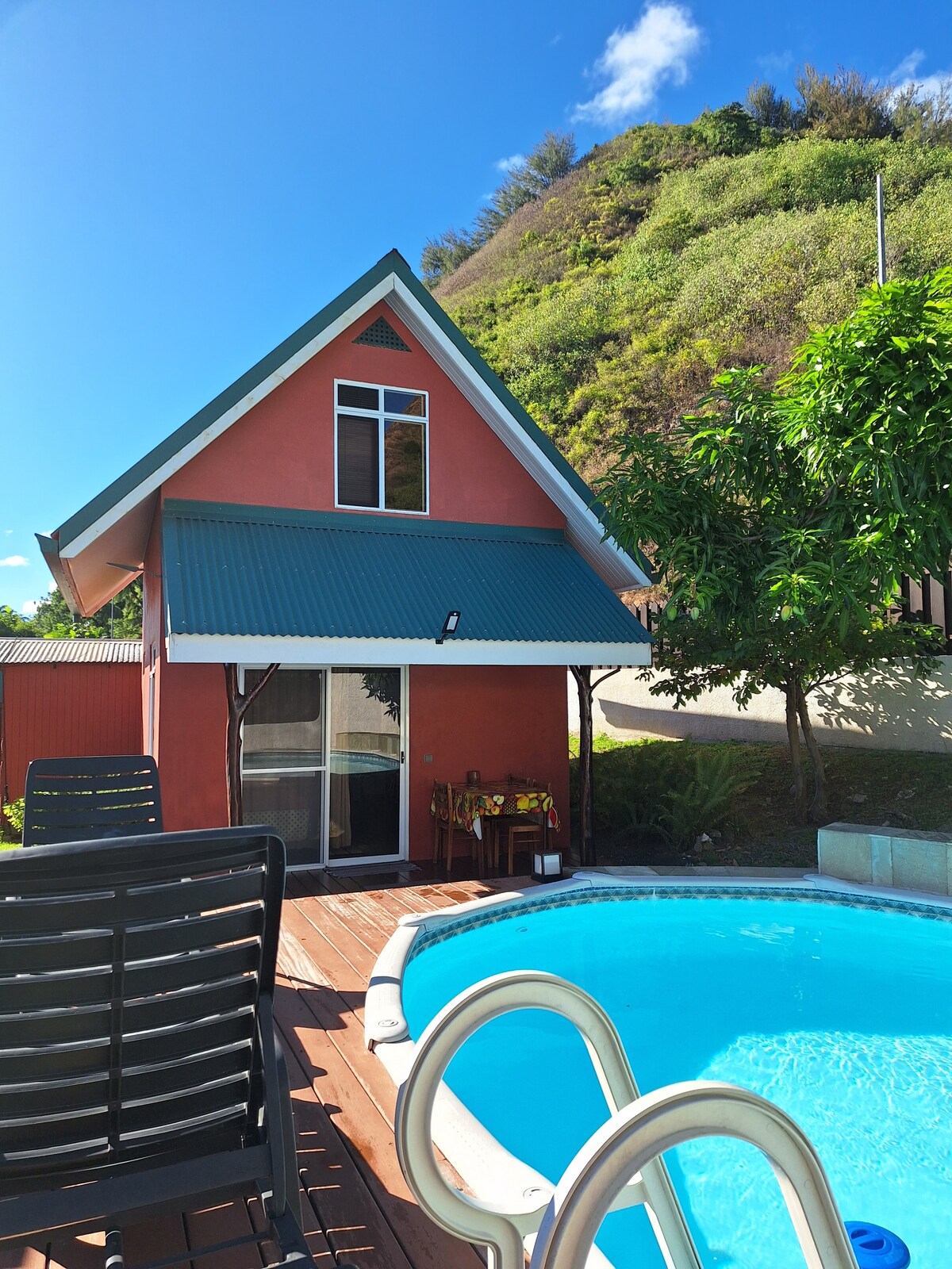 A charming two-story house with a green roof is positioned next to a sparkling blue pool. Wooden decking surrounds the pool, and dark lounge chairs are arranged for relaxation. Lush greenery provides a backdrop that highlights the tranquil setting.