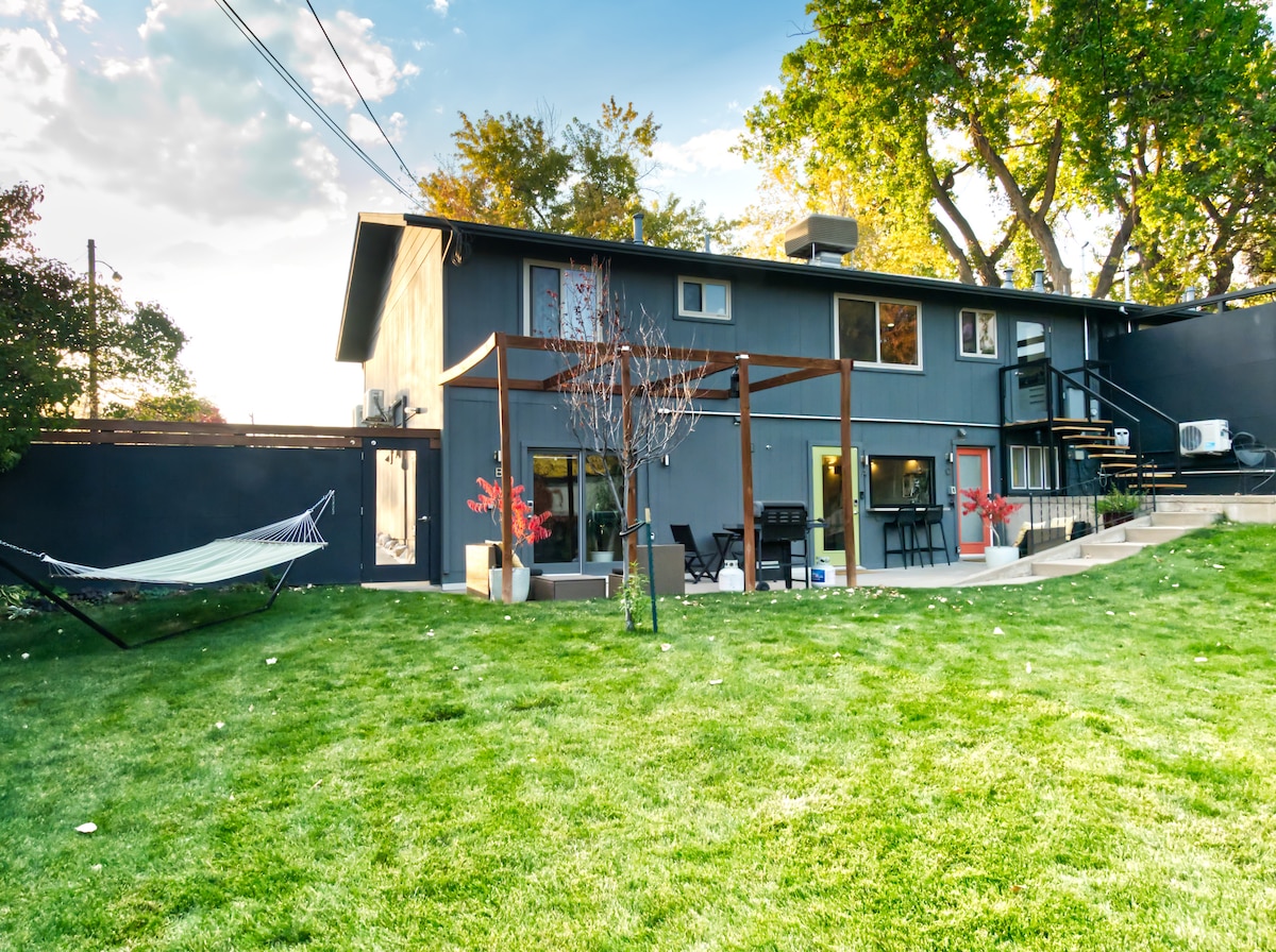 The exterior view showcases a modern two-story home surrounded by a spacious green lawn. A wooden pergola frames a seating area, and a hammock is positioned between two trees. Light filters through the large windows, offering a glimpse of the indoor space.