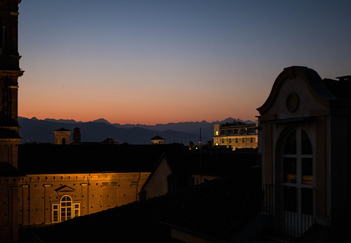 A serene evening view captures the silhouette of distant mountains against a fading sunset sky. The soft gradient from orange to deep blue is complemented by the warm glow of illuminated buildings in the foreground, framed by a window overlooking the tranquil scene.