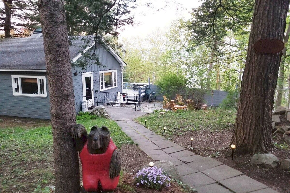 A pathway lined with soft lights leads to a cozy cabin with a gray exterior. A charming bear sculpture in a life jacket stands beside vibrant purple flowers. A spacious deck is visible, surrounded by trees, providing a serene view of the lake.