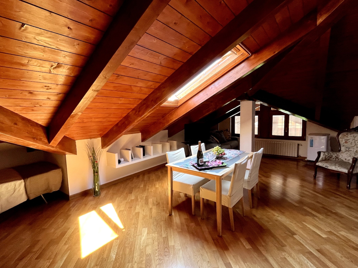 The spacious attic area features sloped wooden ceilings, flooding the room with natural light through a skylight. A dining table is set with breakfast items, complemented by white chairs. A cozy sleeping area with a bed is visible in the background, enhancing the warm atmosphere.