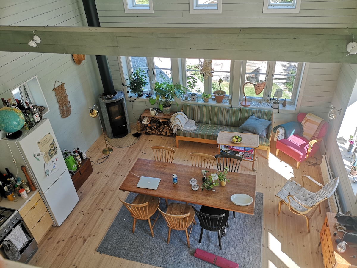 An open-plan living space is shown, featuring a large wooden dining table surrounded by several chairs. Large windows allow ample natural light, highlighting the cozy seating area with cushions and plants. A wood-burning stove is visible, along with elements of an inviting atmosphere.