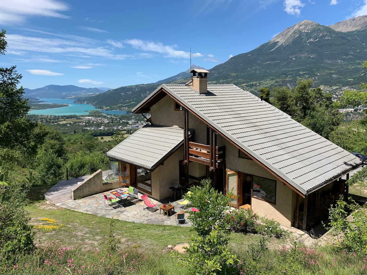 A spacious chalet is showcased, set against a stunning backdrop of mountains and the Serre-Ponçon Lake. Large glass doors provide bright access to the outdoor terrace, lined with colorful seating options. A natural stone pathway leads through lush greenery to the entrance.