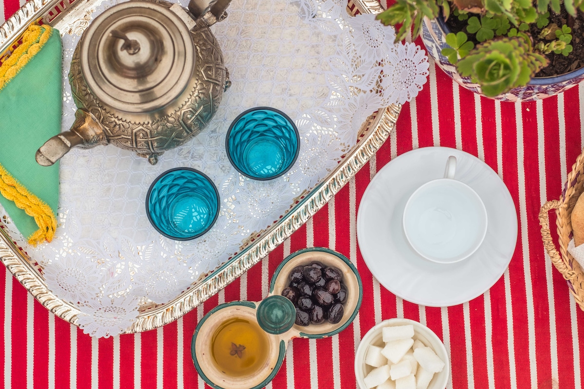 A traditional serving tray is presented with ornate tea tools. Two bright blue glasses accompany a white cup, while an assortment of dates, sugar cubes, and honey is organized in small bowls. The background features a decorative tablecloth with bold red and white stripes.