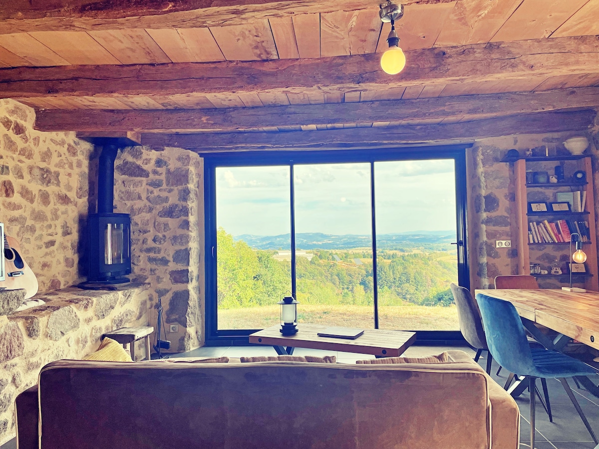 A cozy living area features exposed wooden beams and a stone wall, providing a rustic charm. Large glass doors offer panoramic views of the valley. A comfortable sofa and wooden dining table accommodate relaxation and meals, while a wood stove adds warmth to the space.