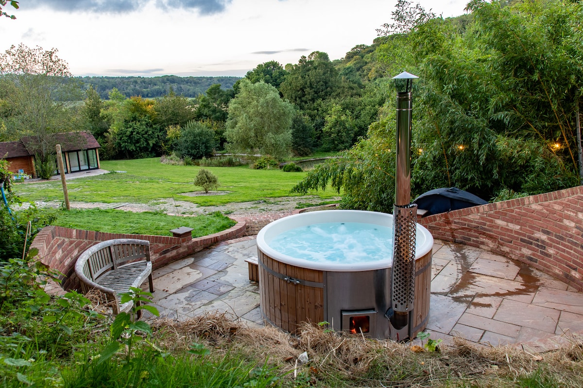 A wood-fired hot tub is positioned on a stone patio, surrounded by lush greenery and scenic hills. A chimney extends from the center, and seating is provided nearby. The inviting spa setting overlooks a spacious garden, creating a tranquil outdoor retreat.