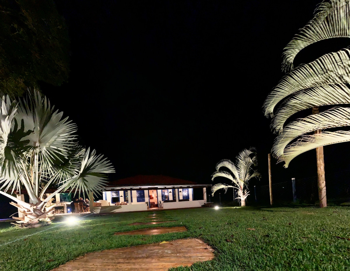 A colonial-style house is illuminated at night, showcasing large windows that provide a glimpse of the cozy interior. The pathway of wooden planks leads through a well-maintained lawn, bordered by tall palm trees, creating an inviting outdoor atmosphere.