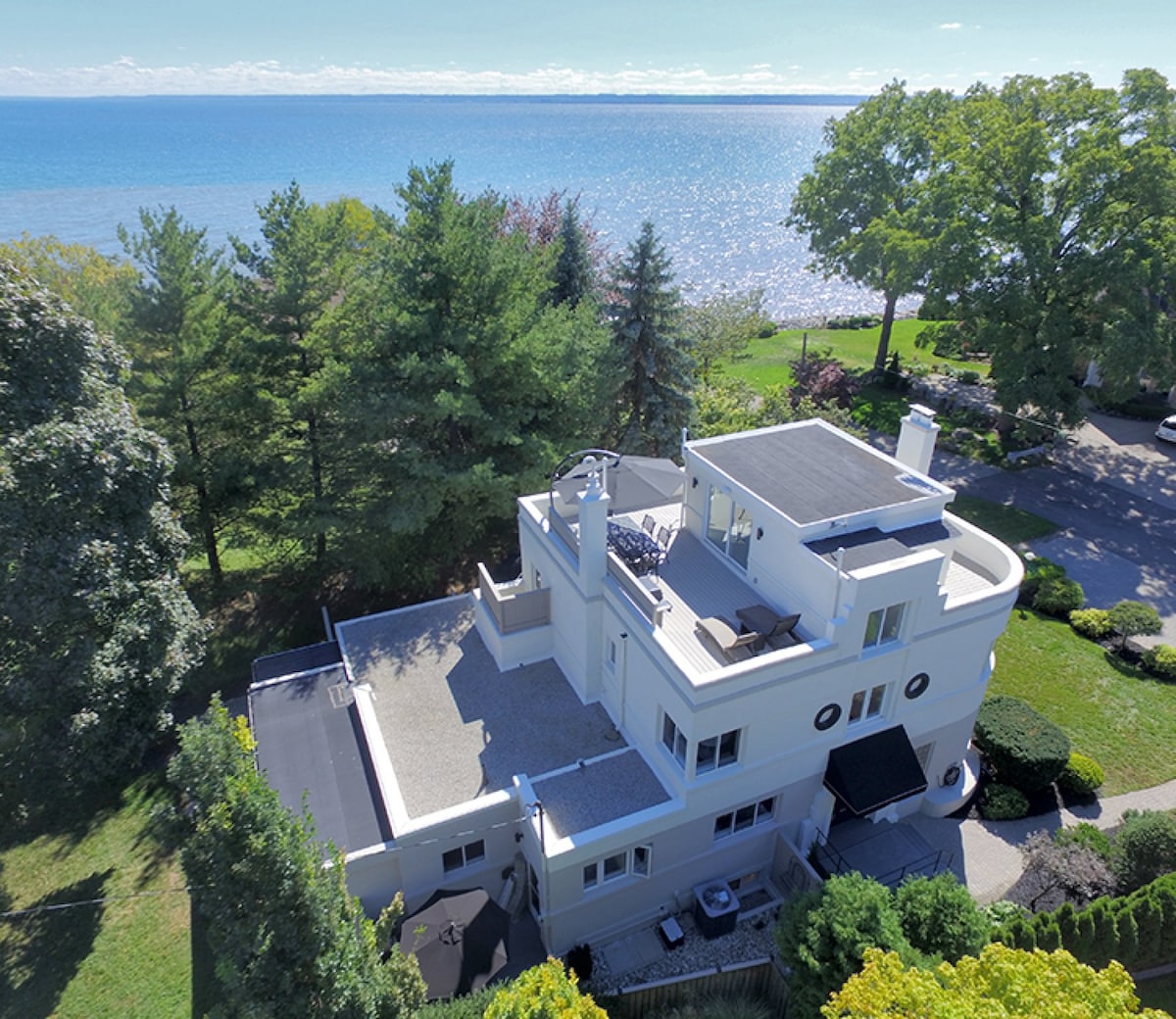 An aerial view of a top-floor apartment is displayed, featuring a rooftop terrace and scenic grounds. The tranquil Lake Ontario glistens in the background, surrounded by trees. The structure showcases modern architectural elements, inviting nature into the living space.