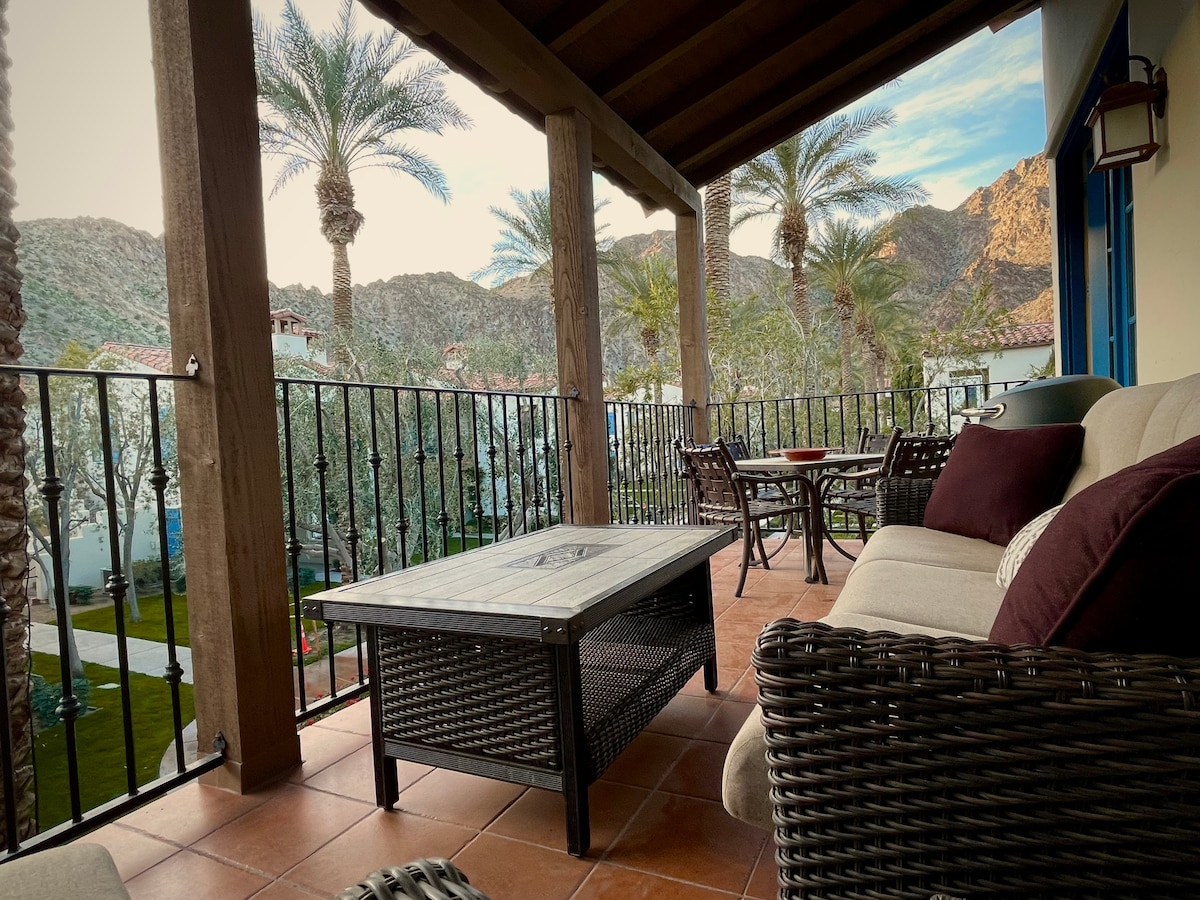 An outdoor balcony features comfortable seating options, including a cushioned couch and armchair with a matching coffee table. Palm trees and mountains create a serene backdrop, enhancing the tranquil atmosphere. The tiled floor adds a warm touch to the inviting space.