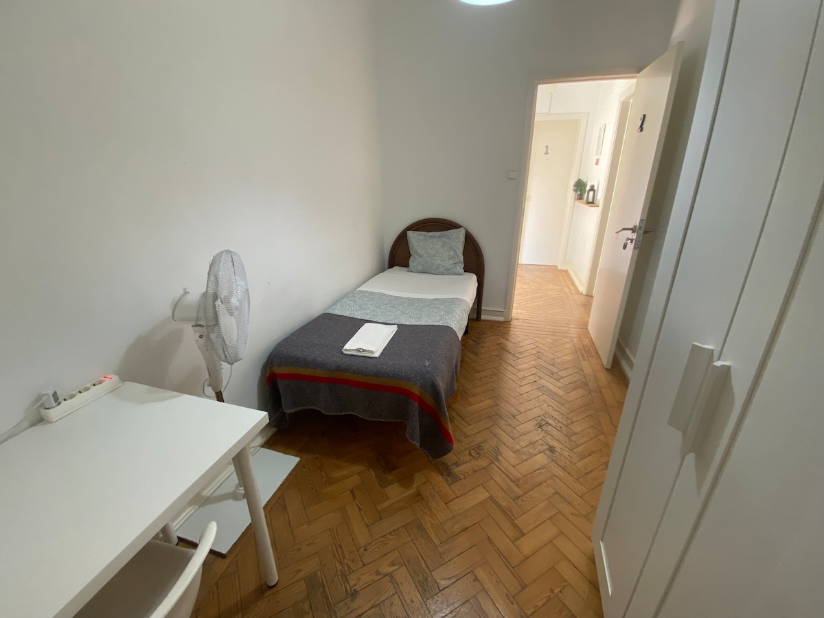 A bright room features a single bed with a neatly arranged gray blanket and a white towel. A fan is positioned nearby, and a simple white desk sits next to the bed. A door leading to the hallway is visible, along with light-colored walls and wooden flooring.