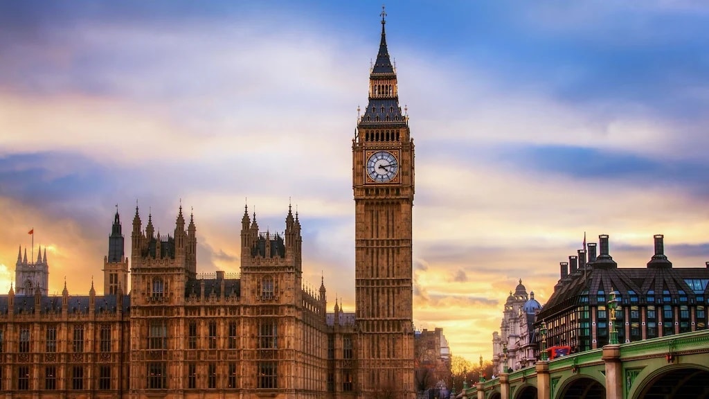 The iconic clock tower, known as Big Ben, stands prominently alongside the Houses of Parliament. The scene features intricate Gothic architecture bathed in warm sunset hues, while the River Thames flows gently beside the historic buildings.