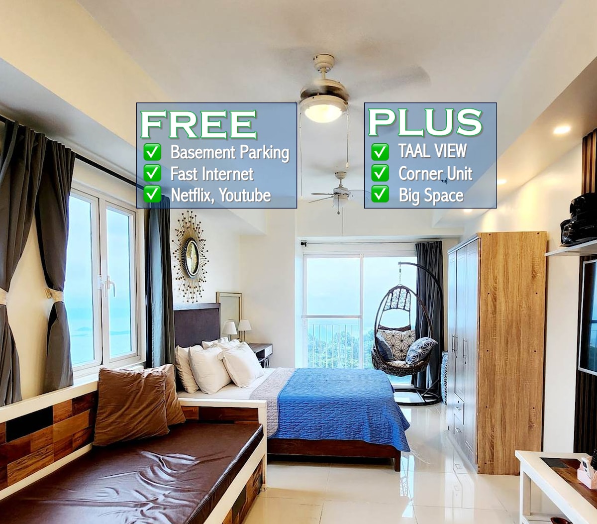 A spacious room features a large bed adorned with white linens and a blue throw. A comfortable seating area includes a hanging chair and modern decor. Natural light floods in through large windows, offering a view of the Taal volcano in the distance.