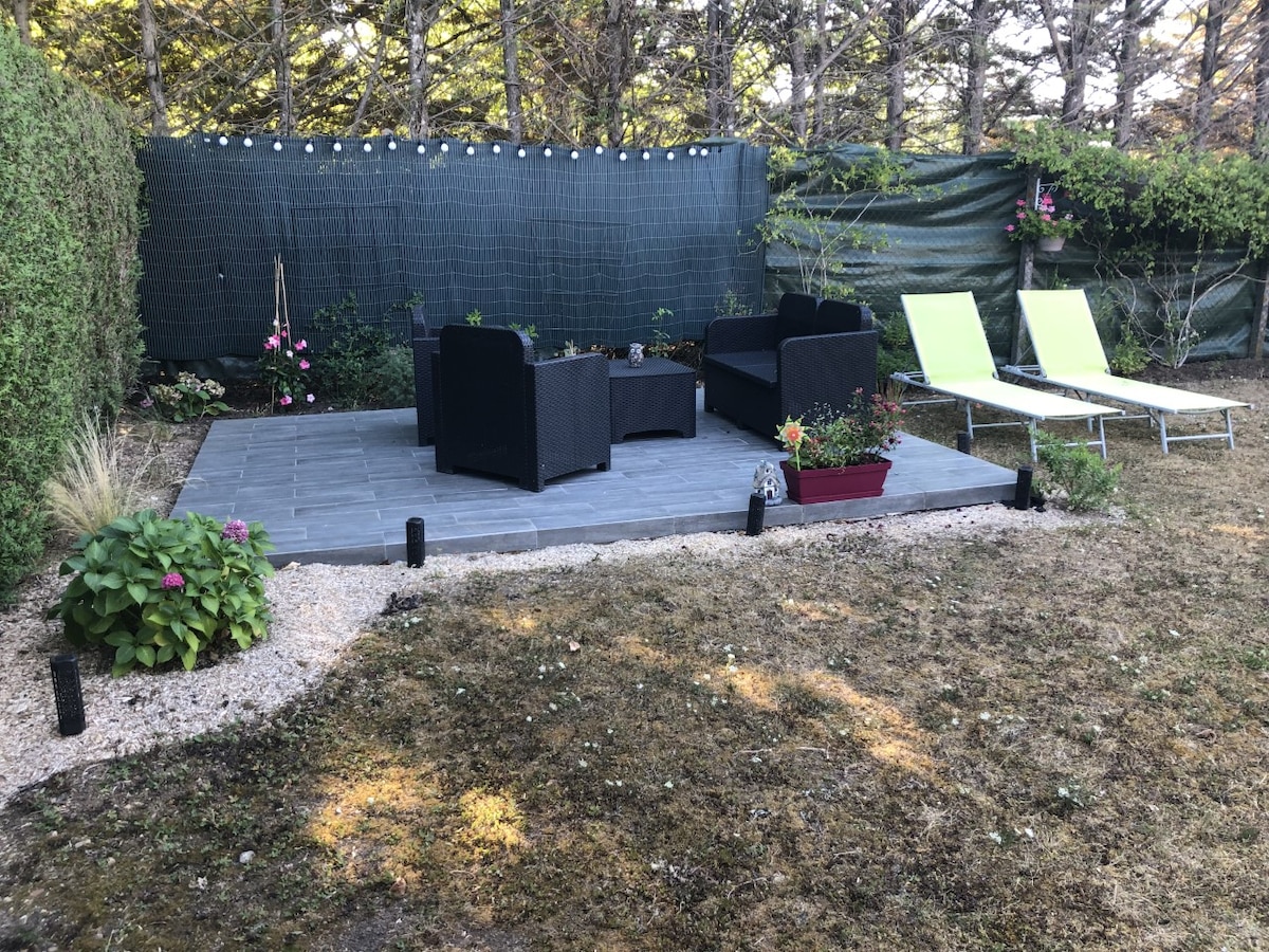 An outdoor patio space is visible, featuring two lounge chairs and a seating area with black wicker furniture. Flower pots and garden accents enhance the setting, while a backdrop of greenery provides privacy. Soft lighting is strung overhead, creating a relaxed atmosphere.