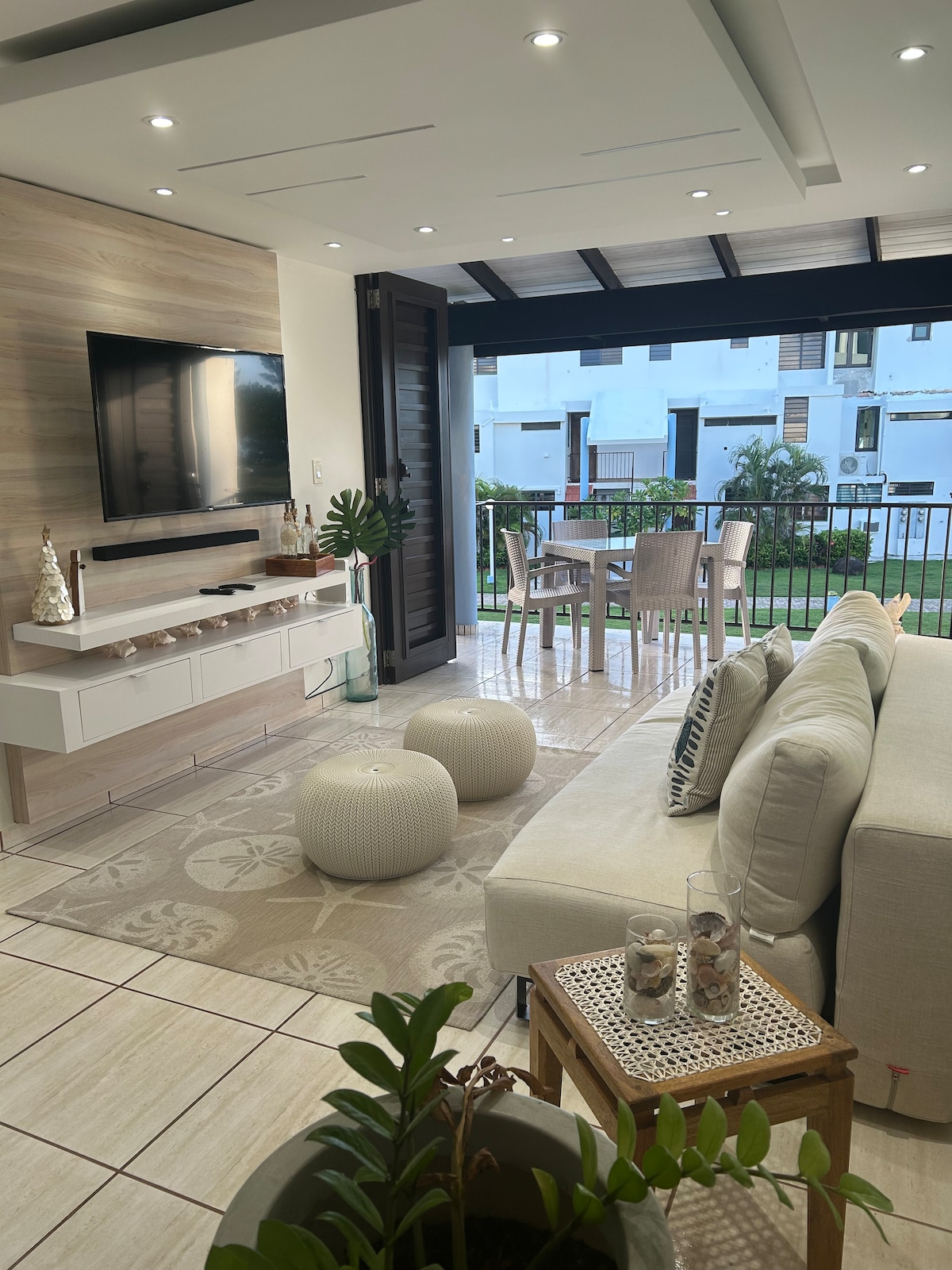 A modern living area is featured with a light-colored sofa and two decorative poufs. A flat-screen TV is mounted on the wooden panel above a sleek media console. Glass doors lead to an outdoor dining space, with a table and chairs overlooking the green lawns.