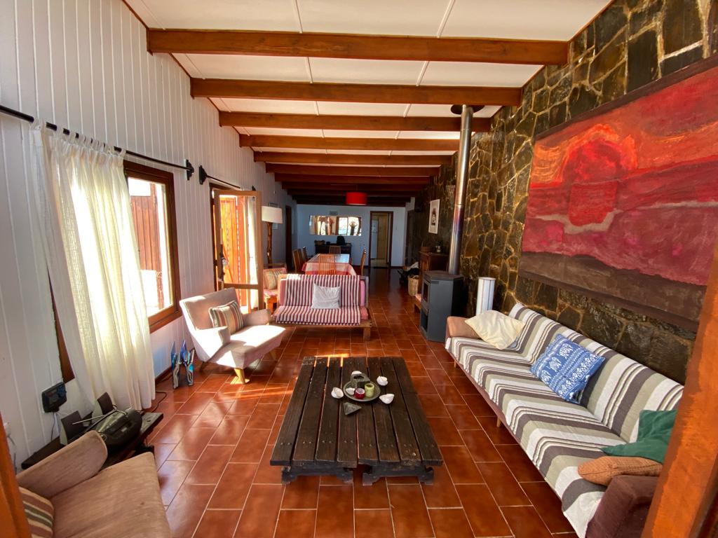 The living area features a blend of seating options, including upholstered chairs and couches with various patterns. Natural light floods through large windows, illuminating the space, while a rustic coffee table sits at the center, adorned with small decorative items. Stone walls provide a contrasting texture.