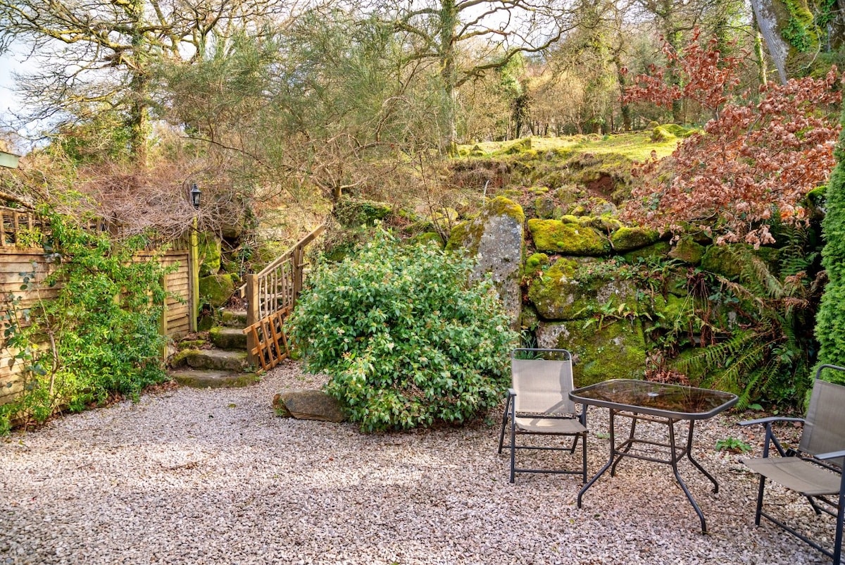 A private gravelled courtyard is framed by lush greenery and moss-covered rocks. Two chairs and a small table are positioned to one side, providing a peaceful spot to relax. A wooden path leads to the steps, enhancing the natural surroundings.