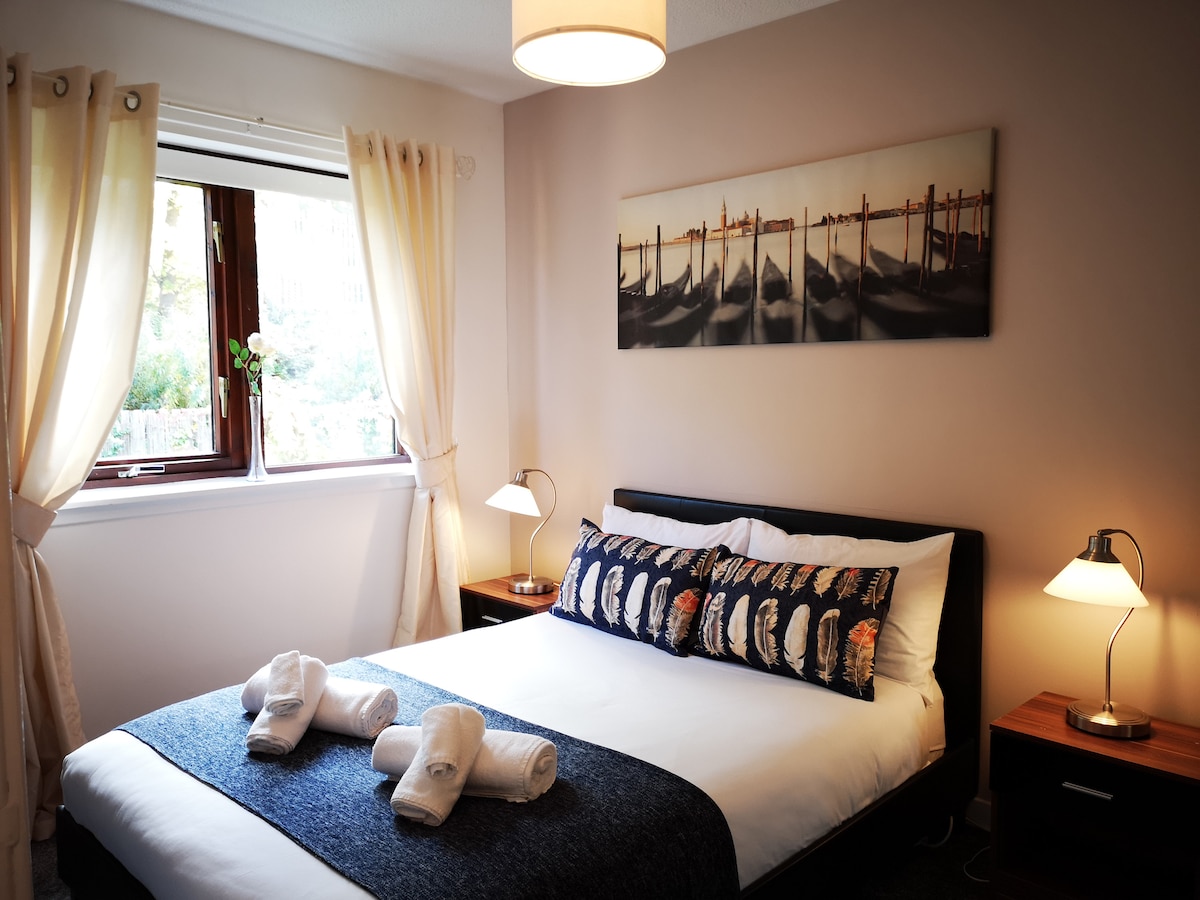 A bright bedroom features a spacious bed with decorative throw pillows and neatly arranged towels. Two bedside tables are positioned on either side, each with a lamp. Natural light filters through a window adorned with cream curtains, illuminating a wall art piece depicting a calm scene.