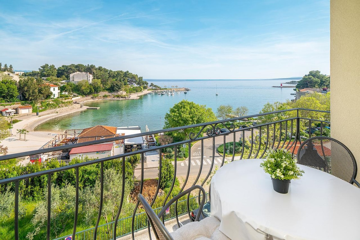 A balcony offers a scenic view of the coastline, with a small beach and boats visible in the water. A round table adorned with a potted plant is surrounded by black chairs, providing a comfortable outdoor seating area.