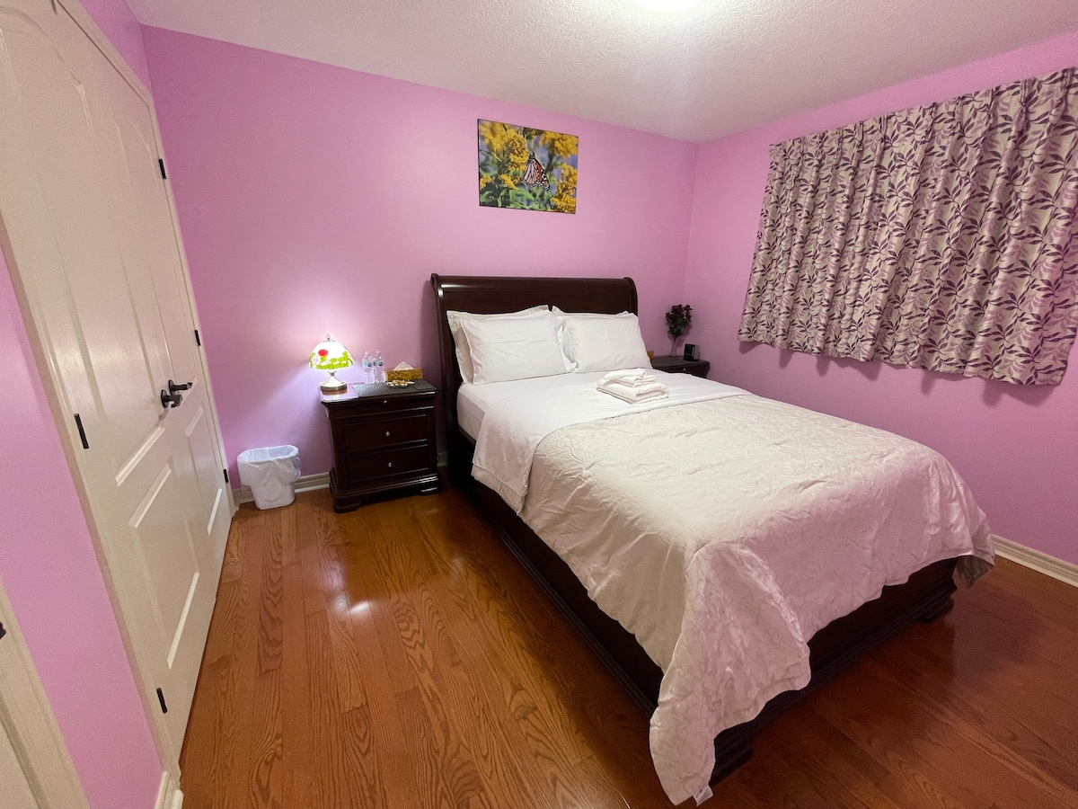 The private room features light pink walls and polished wooden flooring. A spacious bed with fresh linen is positioned against one wall, accompanied by a nightstand with a lamp. Light enters through a curtain-draped window, and a piece of wall art adds a touch of color.