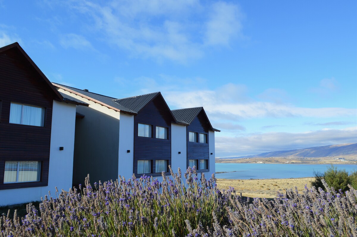 Two multi-story buildings are situated near a serene body of water, framed by lavender plants in the foreground. The architecture features triangular roofs and large windows that provide views of the landscape and lake.