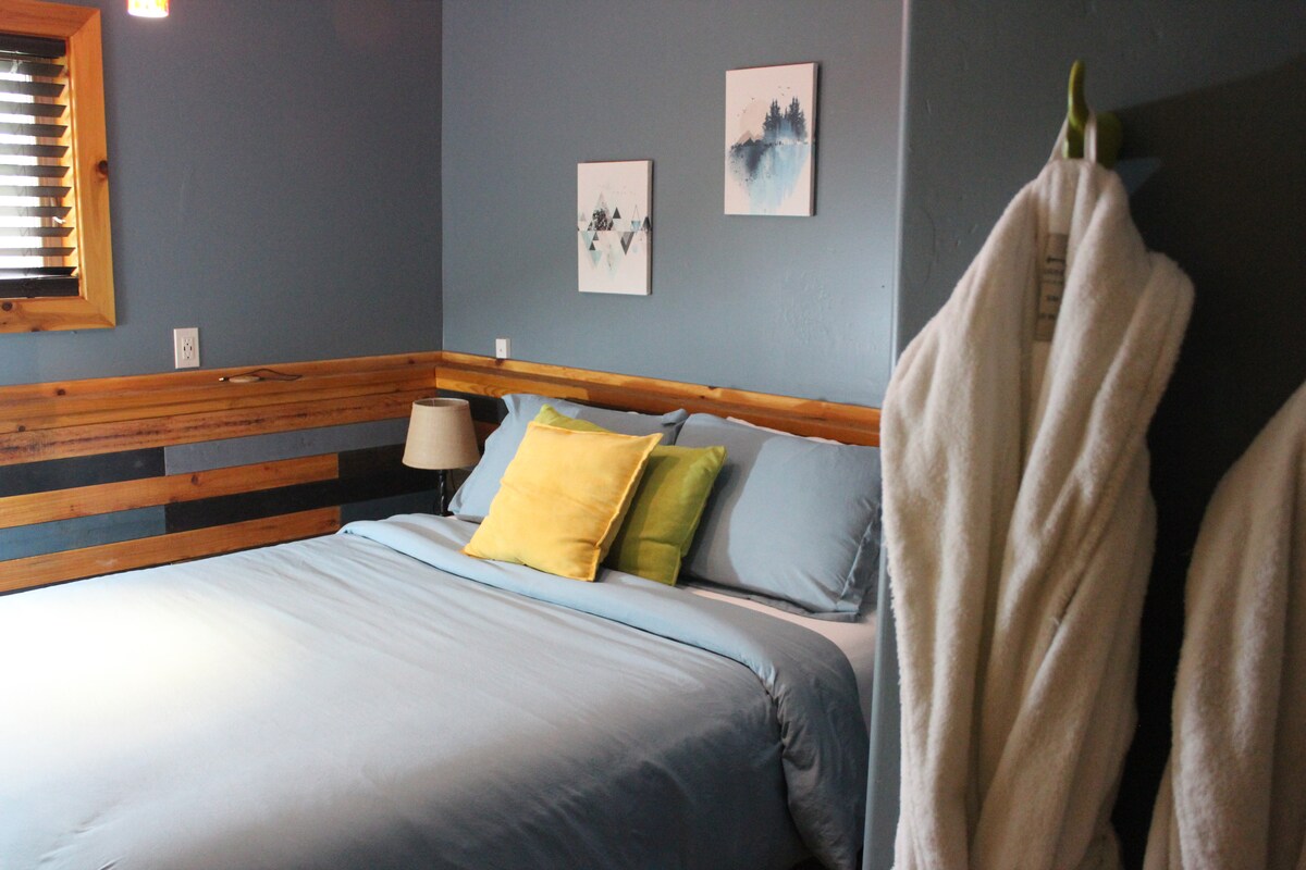 A serene sleeping area features a spacious bed adorned with soft blue bedding and accent pillows in yellow and green. Artwork hangs on the wall, complementing the natural wood paneling. White robes are seen hanging nearby, adding to the welcoming ambiance.