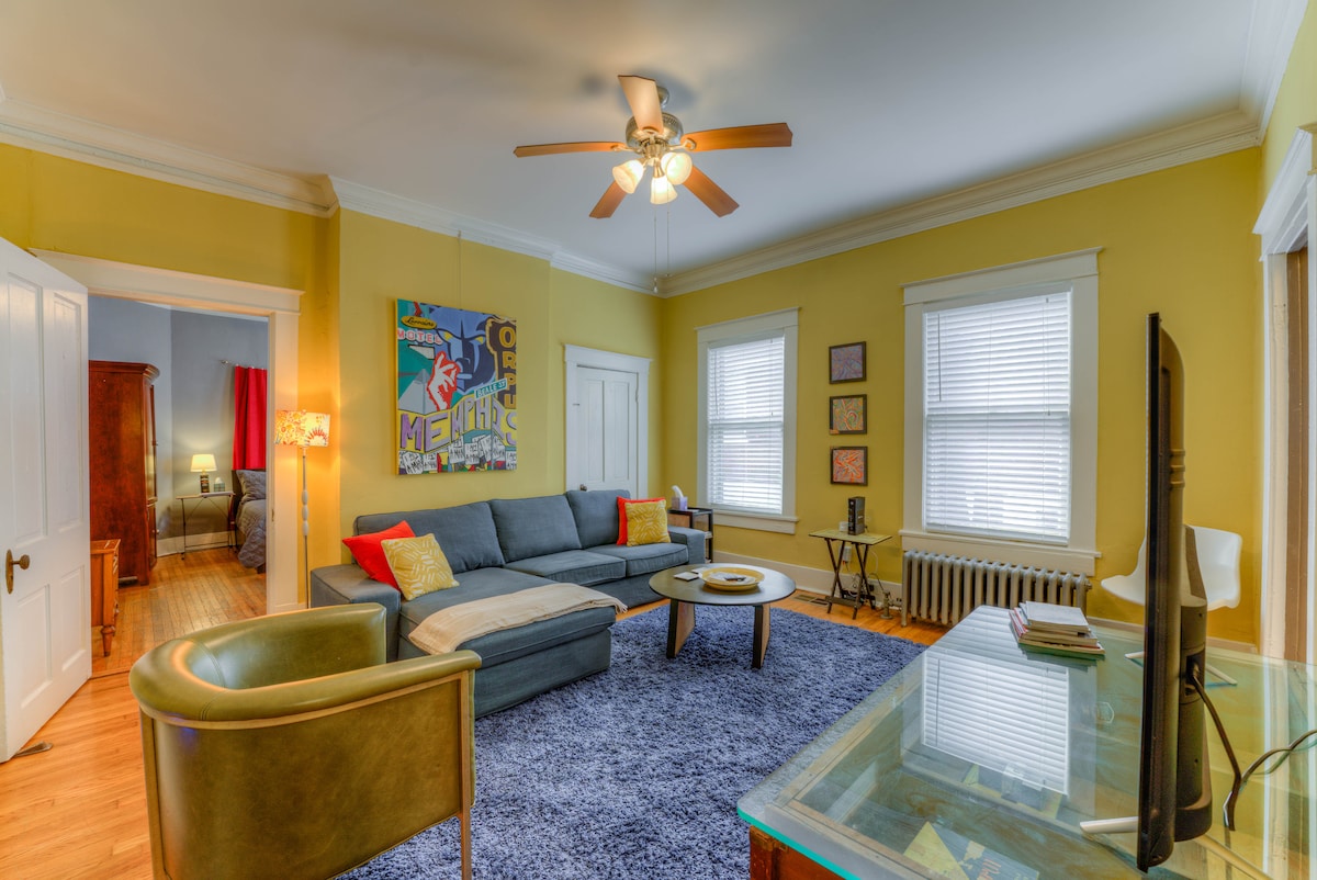 A living room space features a large gray sectional sofa, yellow walls, and decorative artwork. A ceiling fan adds a functional touch, while a coffee table and a green accent chair complement the seating area. Natural light filters through the three windows, illuminating the room.