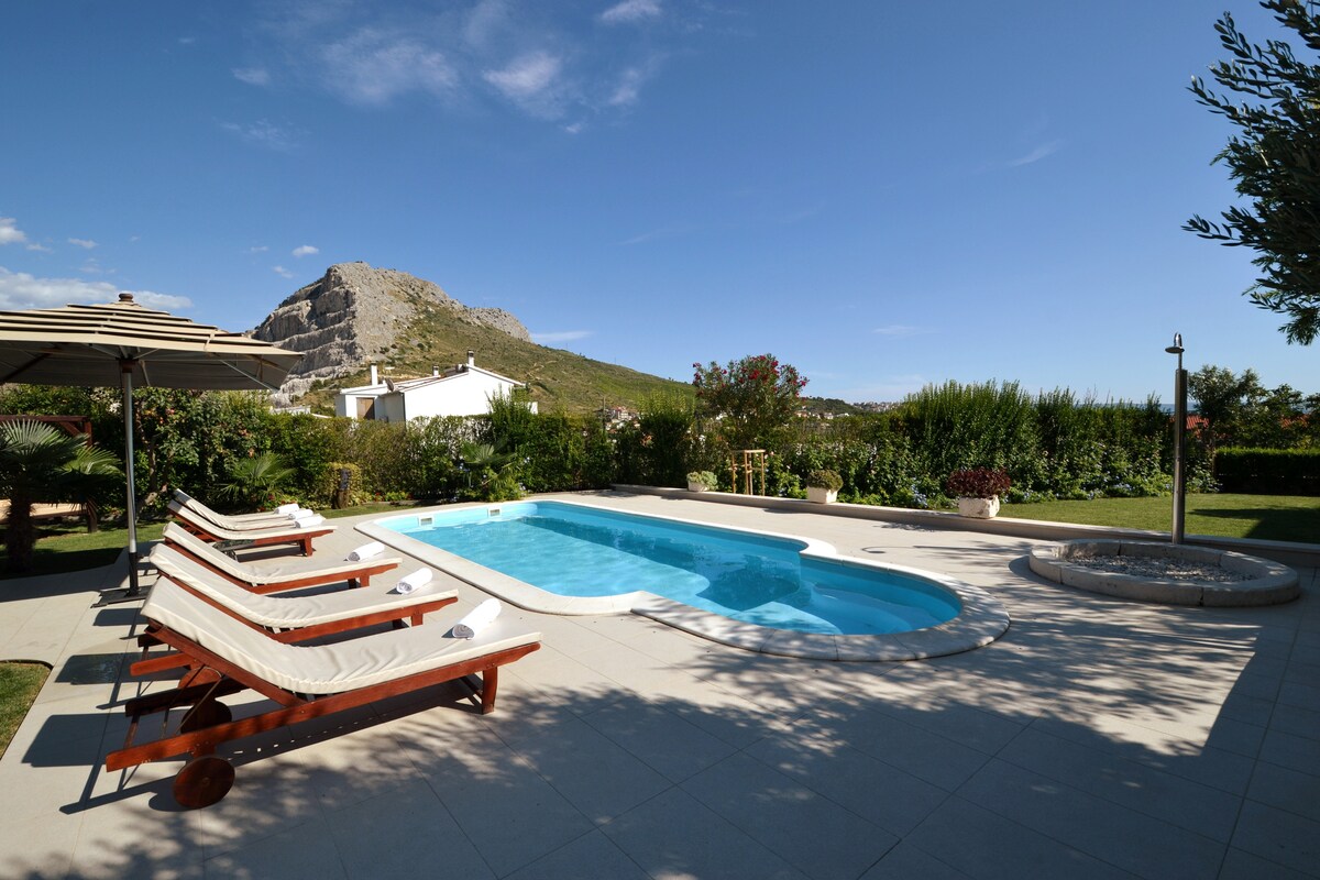 A refreshing pool is surrounded by a spacious patio area, featuring five sunbeds with shades for comfort. Lush greenery lines the property, while the distant mountains frame the serene outdoor setting under a clear blue sky.