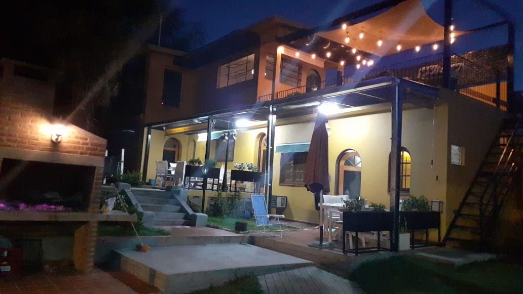 The outdoor area features a spacious patio illuminated by string lights, highlighting comfortable seating arrangements. A built-in grill is positioned nearby, and steps lead up to an upper terrace. The building's colorful facade provides a welcoming atmosphere during evening hours.