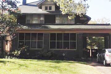 The exterior features a two-story house with a covered porch and multiple windows across the front, allowing natural light to enter. Lush greenery surrounds the base, enhancing the inviting appearance of the residence.