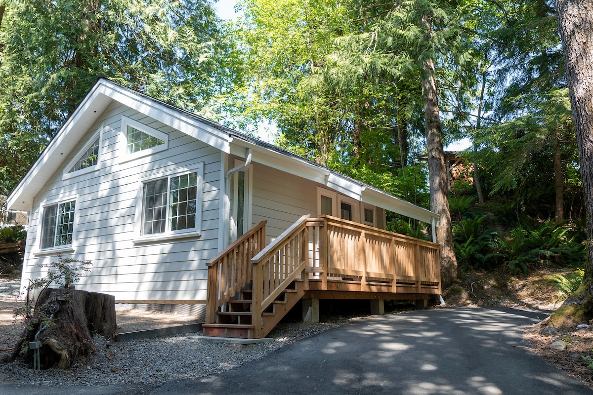 A single-story structure is set amidst tall trees, featuring a wooden ramp leading to a covered entrance. Large windows allow natural light to fill the interior, while a spacious driveway offers easy access and parking.