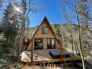The exterior of a renovated A-frame cabin is presented, surrounded by trees. A large deck with a hot tub is visible at the forefront. Large glass doors invite natural light into the interior, while the sloped roof adds to the distinctive architectural style.