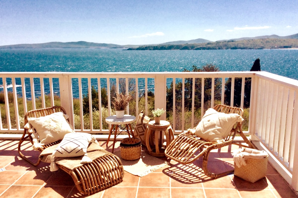 A coastal balcony features two wicker lounge chairs and a small table, set against a stunning view of the blue water and distant hills. Decorative cushions are arranged on the chairs, enhancing comfort, while a woven basket and a small potted plant add texture.