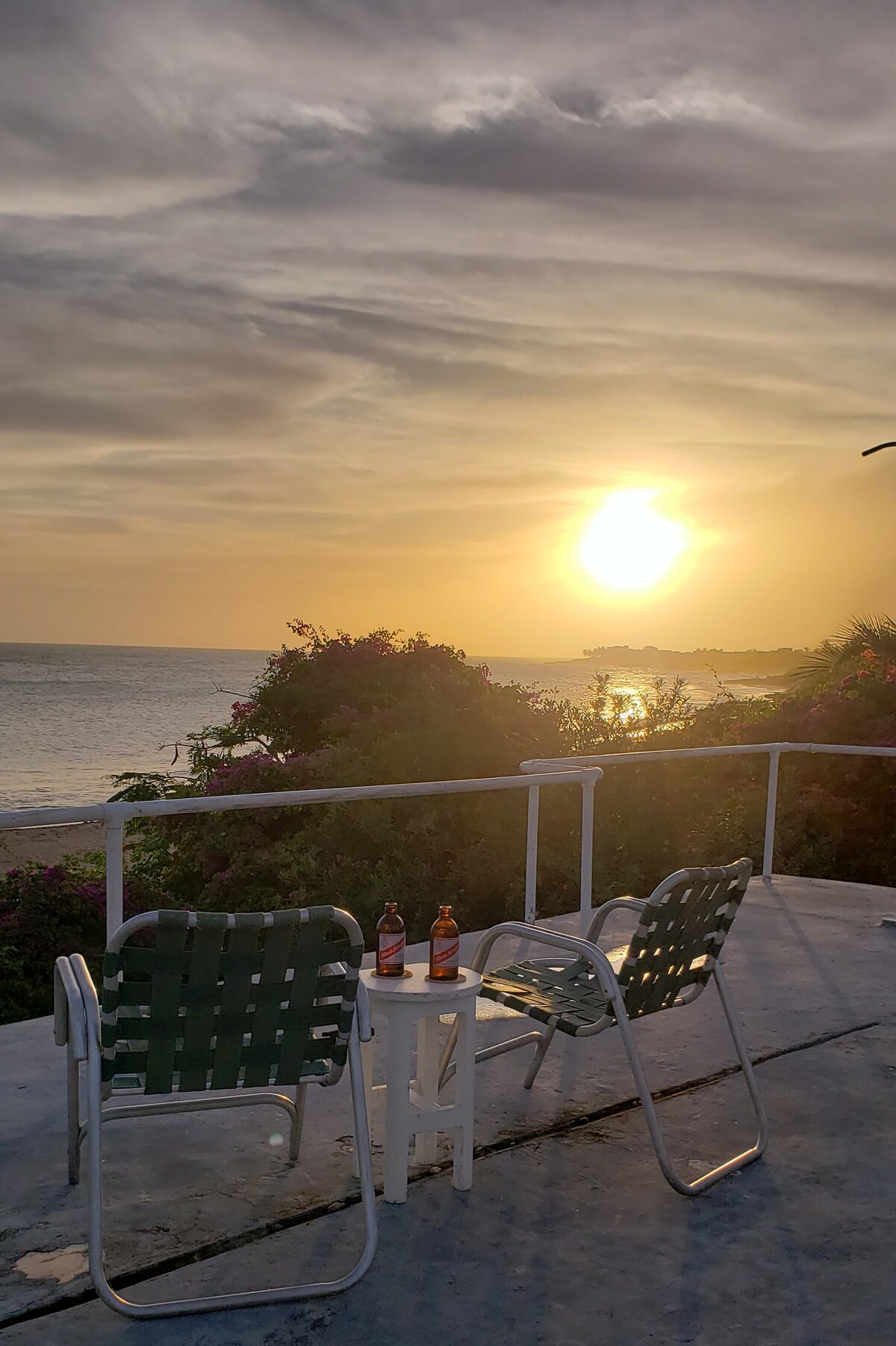 A serene outdoor space features two green patio chairs positioned around a small table. Sunset hues fill the sky as the sun descends, casting a warm glow over the nearby sea. Lush vegetation enhances the tranquil setting.