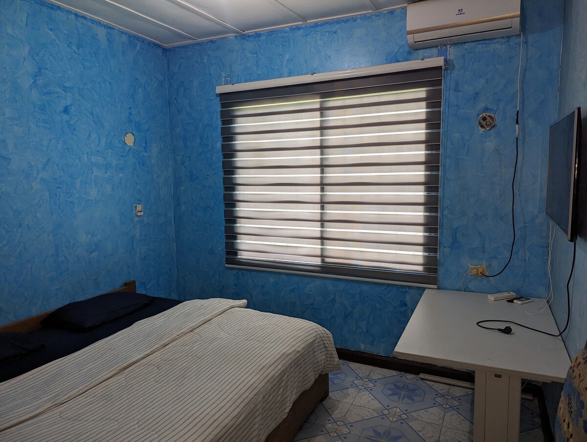 A relaxed bedroom is painted in a soft blue hue, featuring a spacious bed with dark linens. A layered window treatment comprises light-filtering shades and blinds. A simple desk is positioned against the wall, and a wall-mounted television is visible.