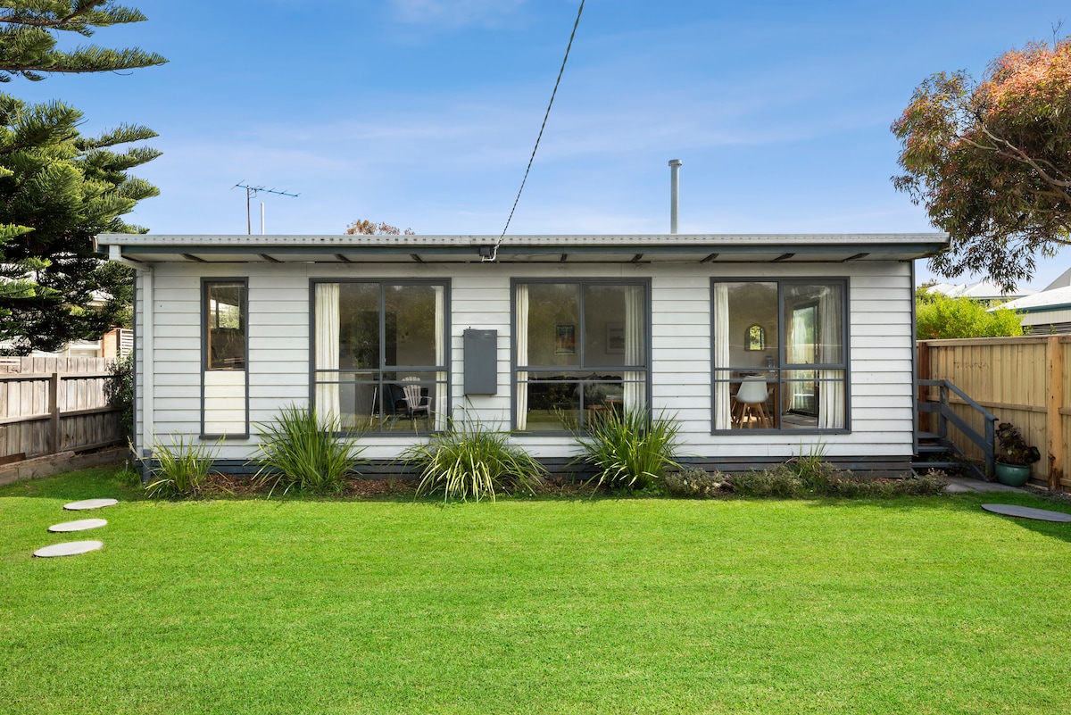 The exterior of Corsair Cottage is showcased, featuring a single-story structure with large windows and a welcoming porch. Lush green grass surrounds the cottage, accented by carefully placed stepping stones leading to the entrance. A peaceful setting is enhanced by nearby trees.