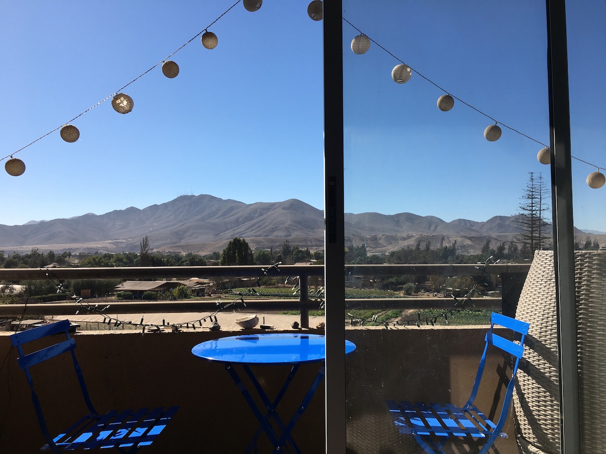 A terrace is presented with a blue round table and two matching chairs, offering a scenic view of distant mountains. Decorative lights hang above, illuminating the space naturally, and a railing adds to the balcony’s openness.