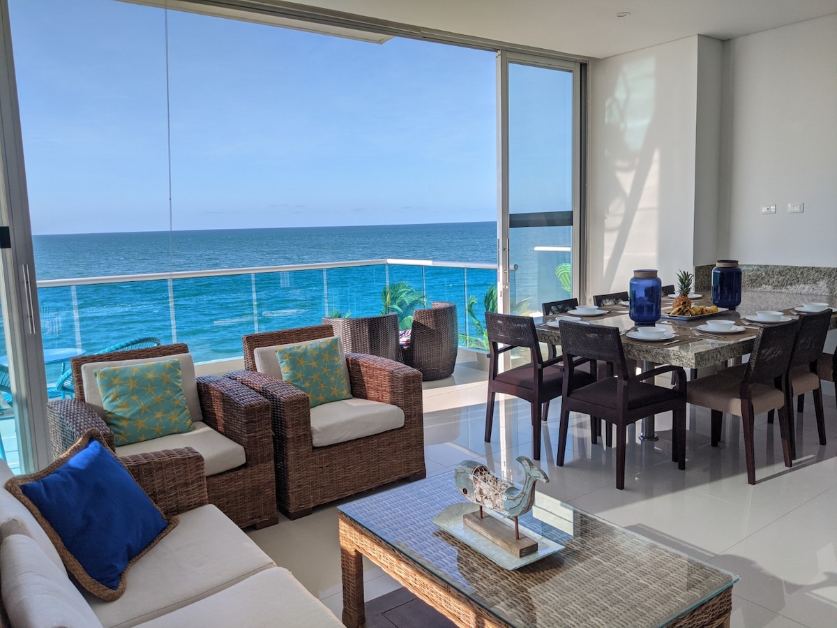 A modern living space features large glass doors that provide an unobstructed view of the ocean. Comfortable seating is arranged around a wooden coffee table, while a dining table accommodates several guests. The decor subtly complements the serene coastal atmosphere.