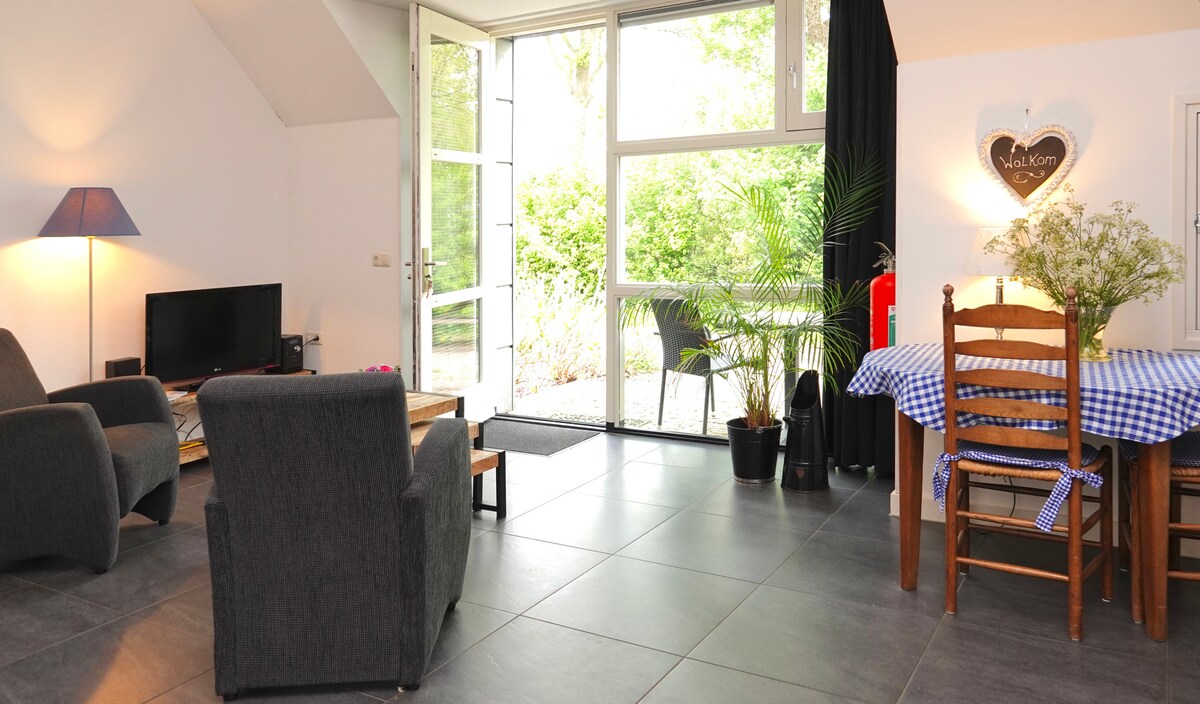 A modern living space showcases a cozy seating area with two dark armchairs and a coffee table. Natural light filters through large glass doors, enhancing the connection to the outdoors. A dining table with a checkered tablecloth and a small indoor plant are visible, contributing to the welcoming environment.