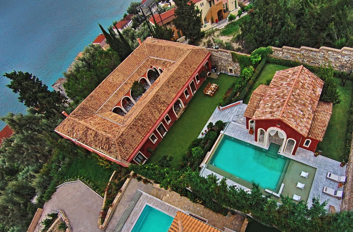 An aerial view captures the Villa Veneziano's architectural elegance, surrounded by lush greenery. The large swimming pool is situated beside a charming pool house, with red-tiled roofs complementing the serene landscape. The distant coastline is visible, enhancing the picturesque setting.
