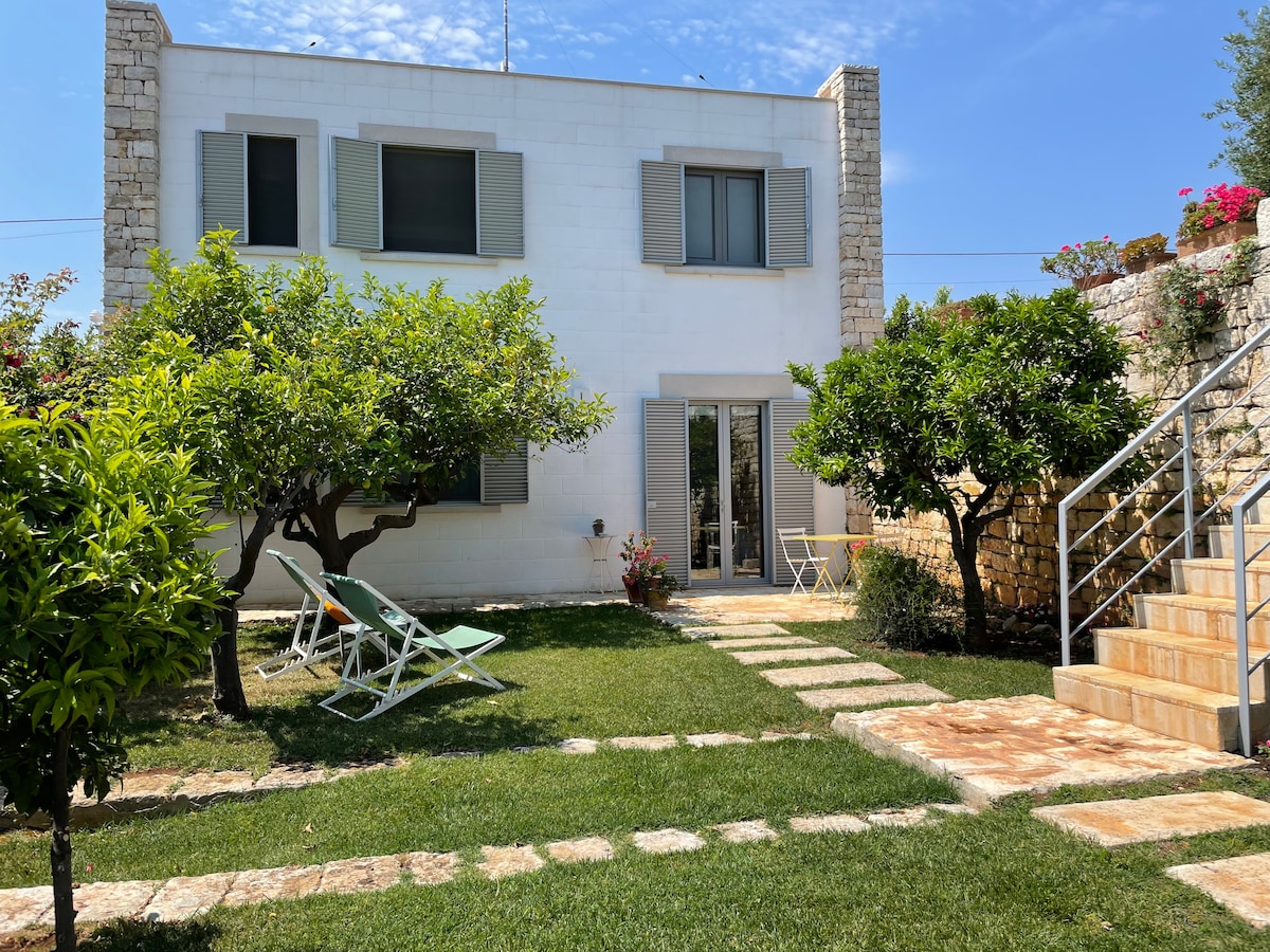 The exterior of a two-story white house is visible, flanked by greenery and flowering plants. A well-maintained lawn features stone pathways leading to a pair of loungers and a small outdoor dining set. The building is accented by large windows dressed in shutters.