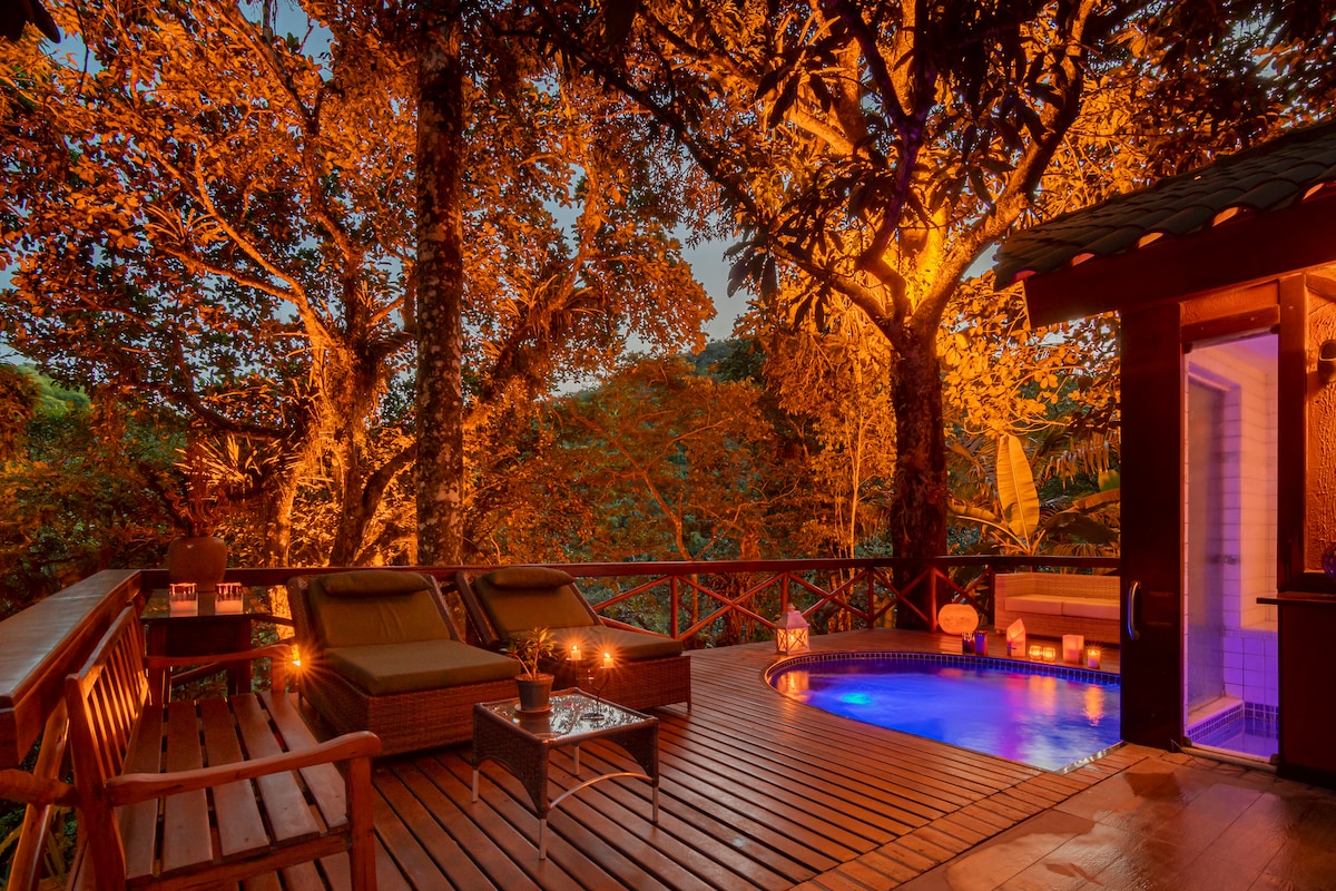 A warm-lit outdoor area features a spa-like pool, surrounded by lush trees. Comfortable lounge chairs are placed on a wooden deck, while soft lighting creates a calming ambiance. A small table holds decorative items, enhancing the tranquil experience of this inviting space.