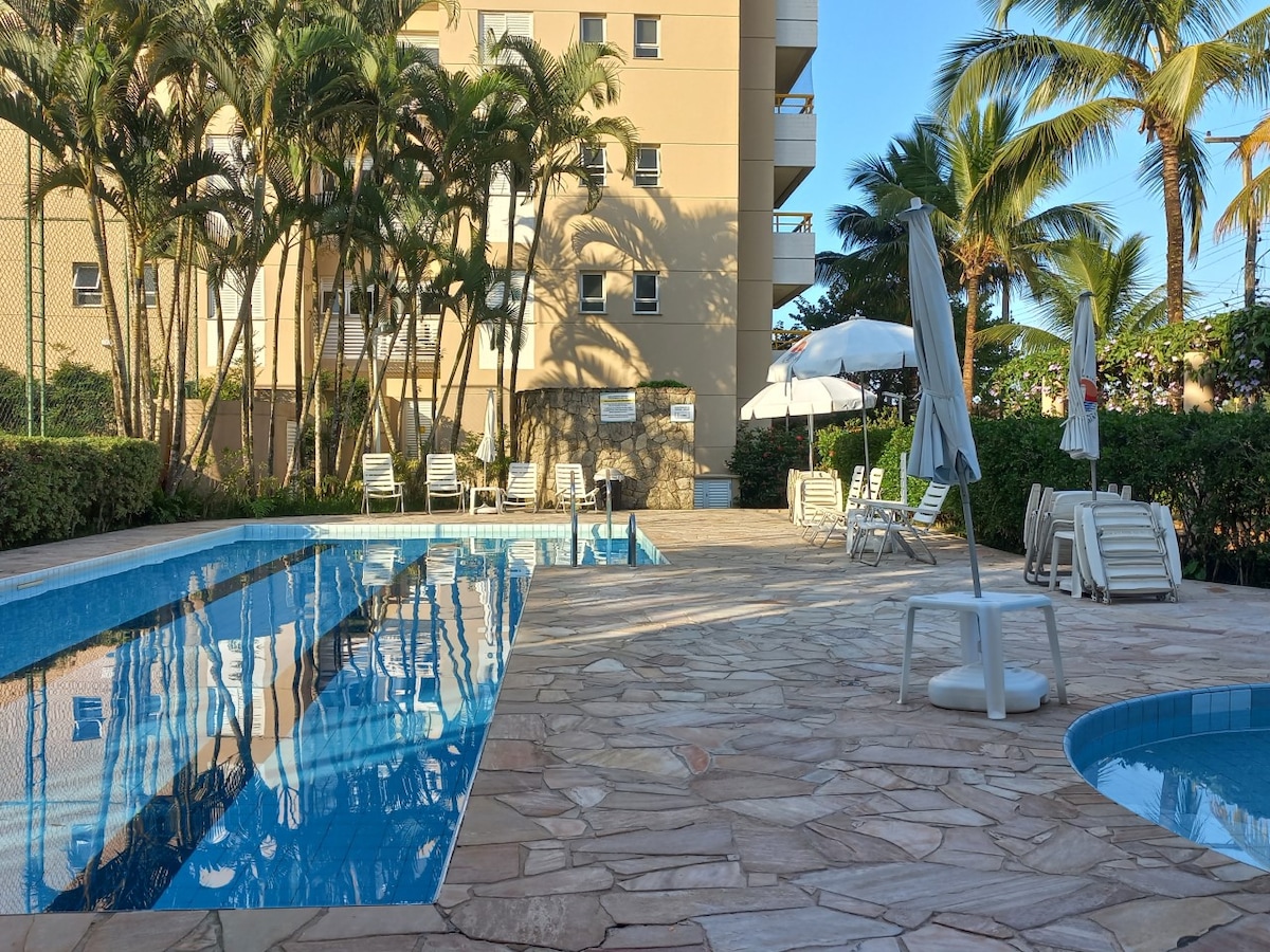A tranquil pool area is surrounded by palm trees and well-maintained landscaping. Sun loungers are arranged on the poolside patio, alongside a shaded table and umbrellas. The water's surface reflects the clear sky and nearby buildings, creating a serene atmosphere.