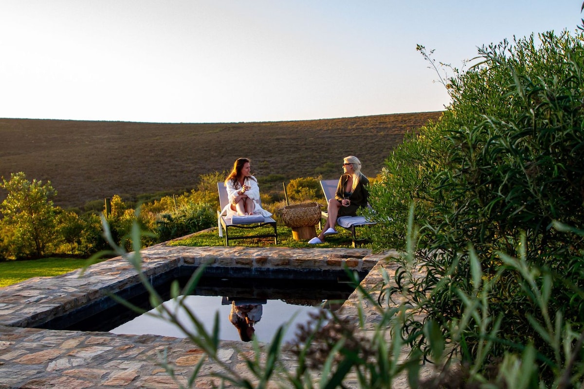 Two guests are seated on comfortable chairs next to a small pool, surrounded by greenery. The landscape features rolling hills in the background, basking in warm sunlight. The setting offers a sense of tranquility and connection to nature.